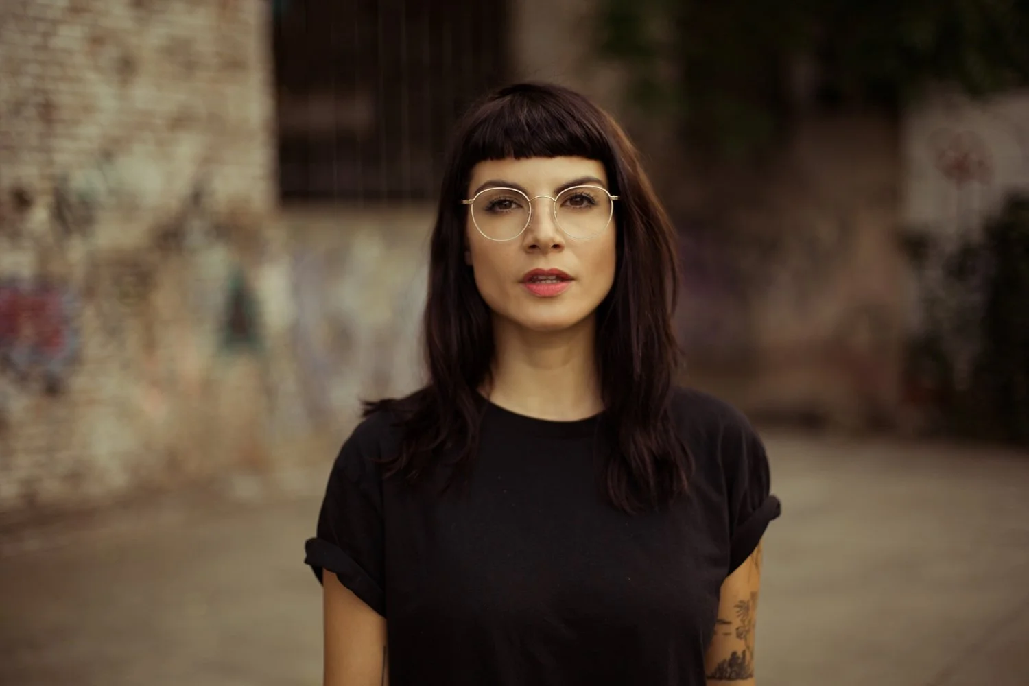 A woman with dark brown hair and bangs, wearing round glasses and a black t-shirt, standing outdoors in front of a blurred brick wall.