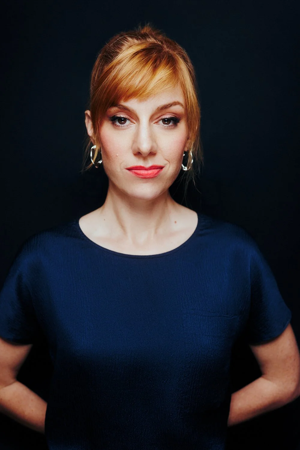 A woman with red hair and light makeup, wearing a dark blue shirt and silver hoop earrings, standing against a black background.