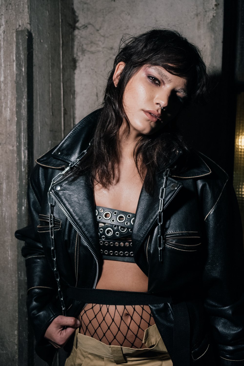 A woman with dark hair and makeup, wearing a black leather jacket, fishnet stockings, and a patterned crop top, standing in an industrial setting.