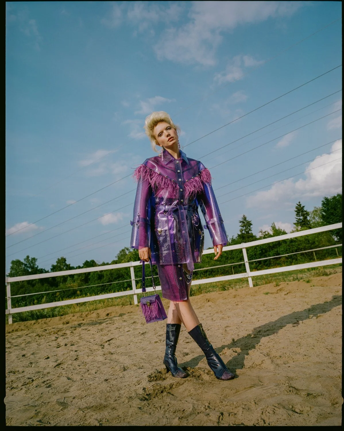 Woman in purple and pink shiny outfit with feather details, holding a matching purple handbag, standing outdoors on sandy ground with a white fence and green trees in the background, under a partly cloudy sky.