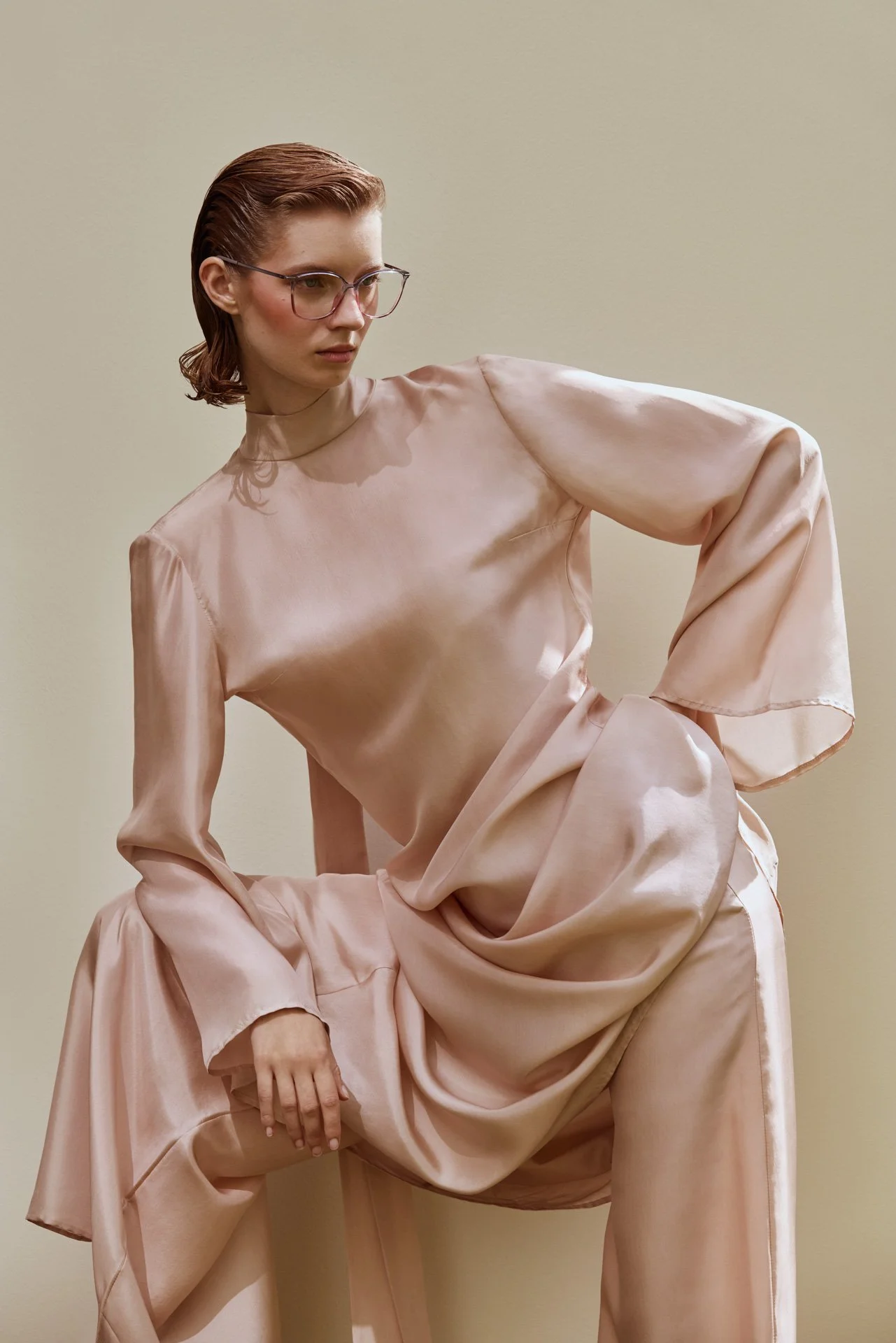 A woman with glasses and short, brown hair poses in a flowing, pale pink satin dress against a plain, beige background.