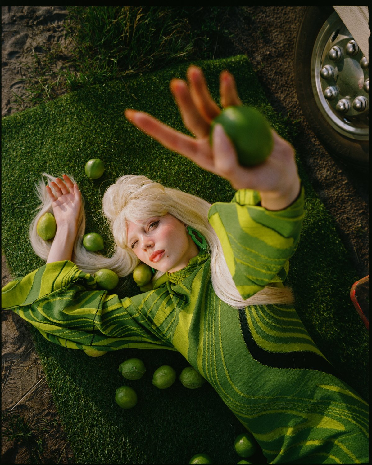 A woman with platinum blonde hair lying on artificial grass surrounded by green limes, wearing a green patterned outfit, holding a lime in her outstretched hand.