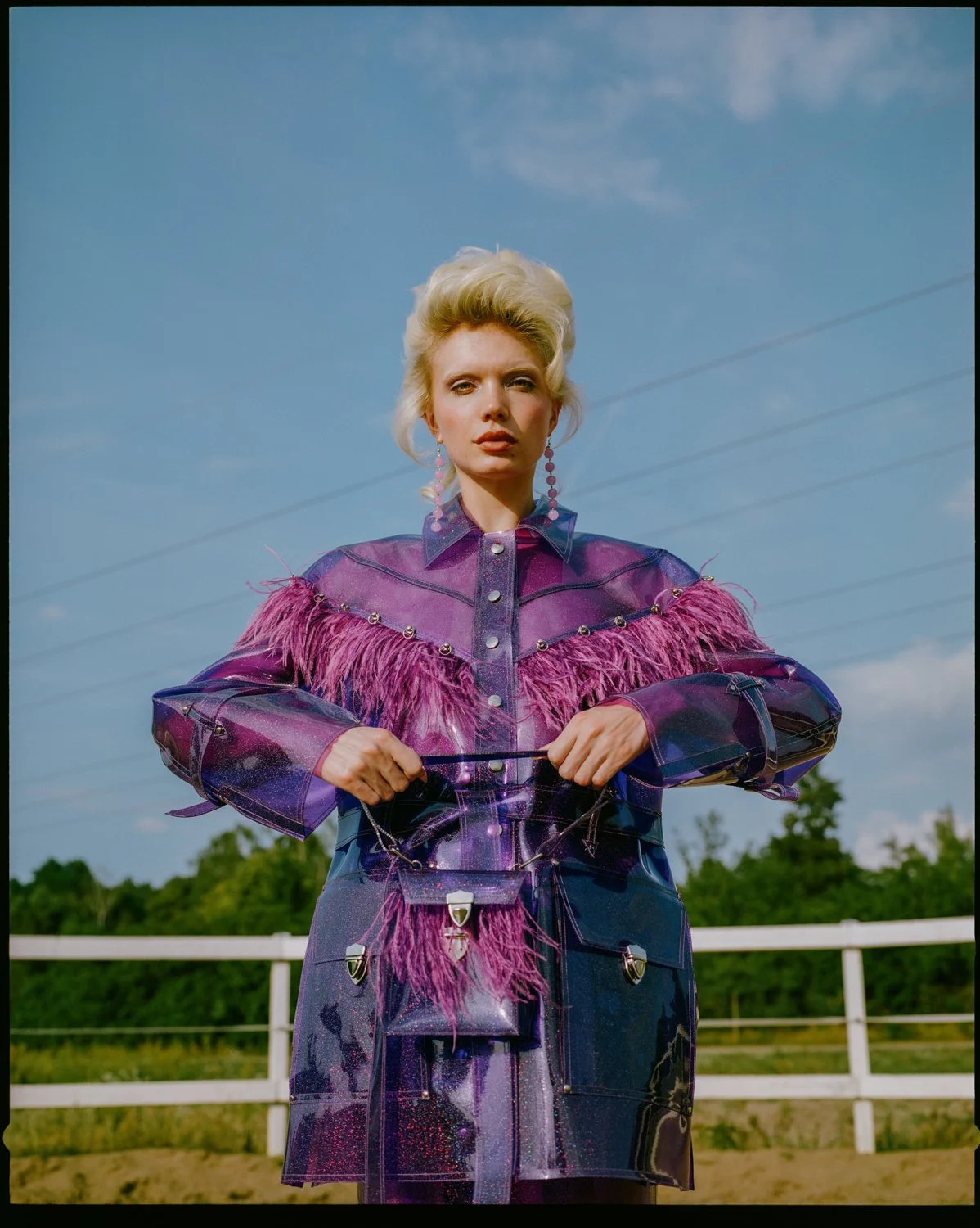 A woman wearing a shiny purple and pink fringed and bejeweled leather outfit, standing outdoors against a blue sky with overhead power lines and green trees in the background.