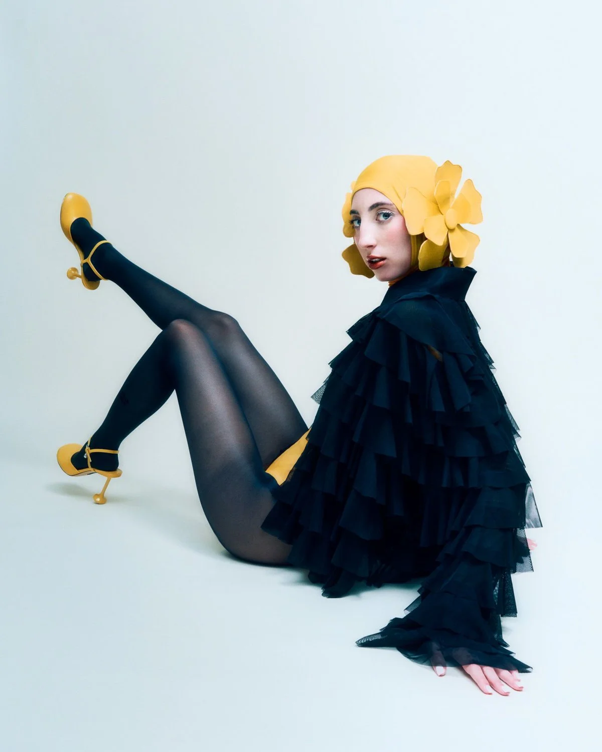 A woman wearing a black layered tulle outfit, yellow high heels with ankle straps, opaque black tights, and a yellow head covering with large decorative yellow flowers, sitting on the floor with one leg raised and the other extended, against a plain 