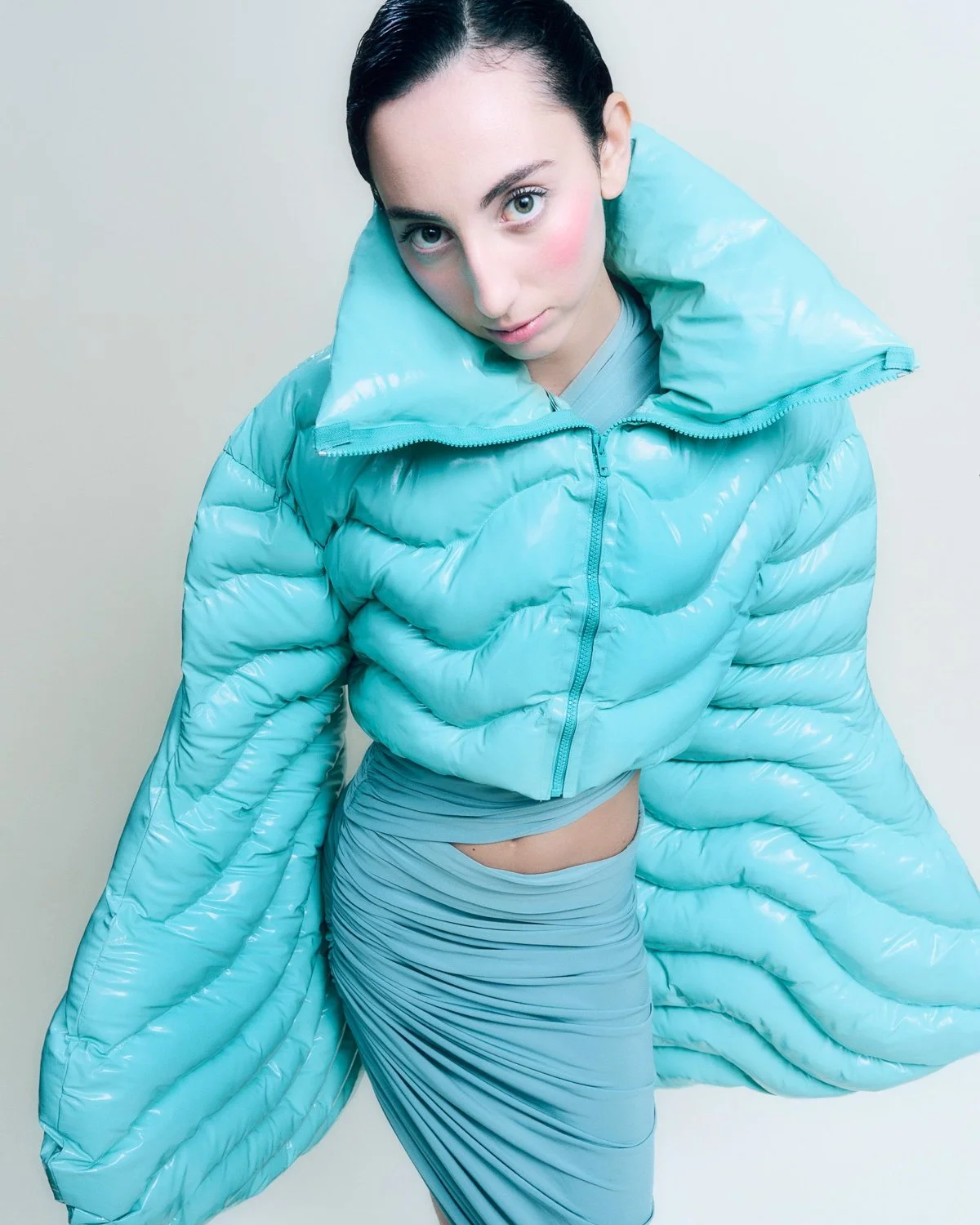 A young woman with dark hair pulled back, wearing a shiny, light blue puffer jacket with a large collar, and a matching fitted skirt. She is looking directly at the camera against a plain, light-colored background.