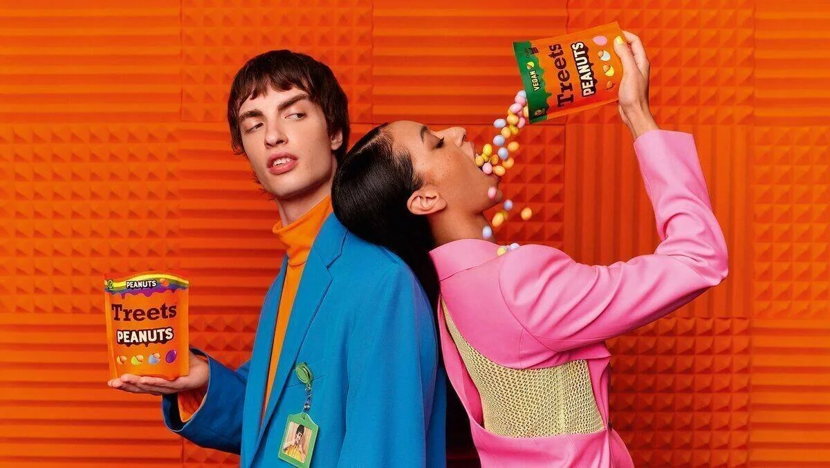 Two young individuals, one male and one female, are posing against an orange textured background. The male is holding a bright orange container labeled "Treets Peanuts" and has a skeptical expression. The female is leaning her head back and pouring m