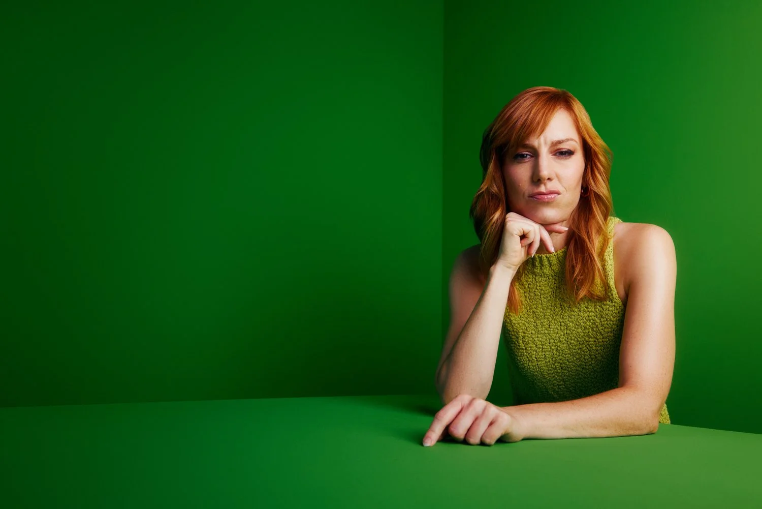 A woman with red hair in a green sleeveless top, resting her chin on her hand, sitting at a green table against a green background.