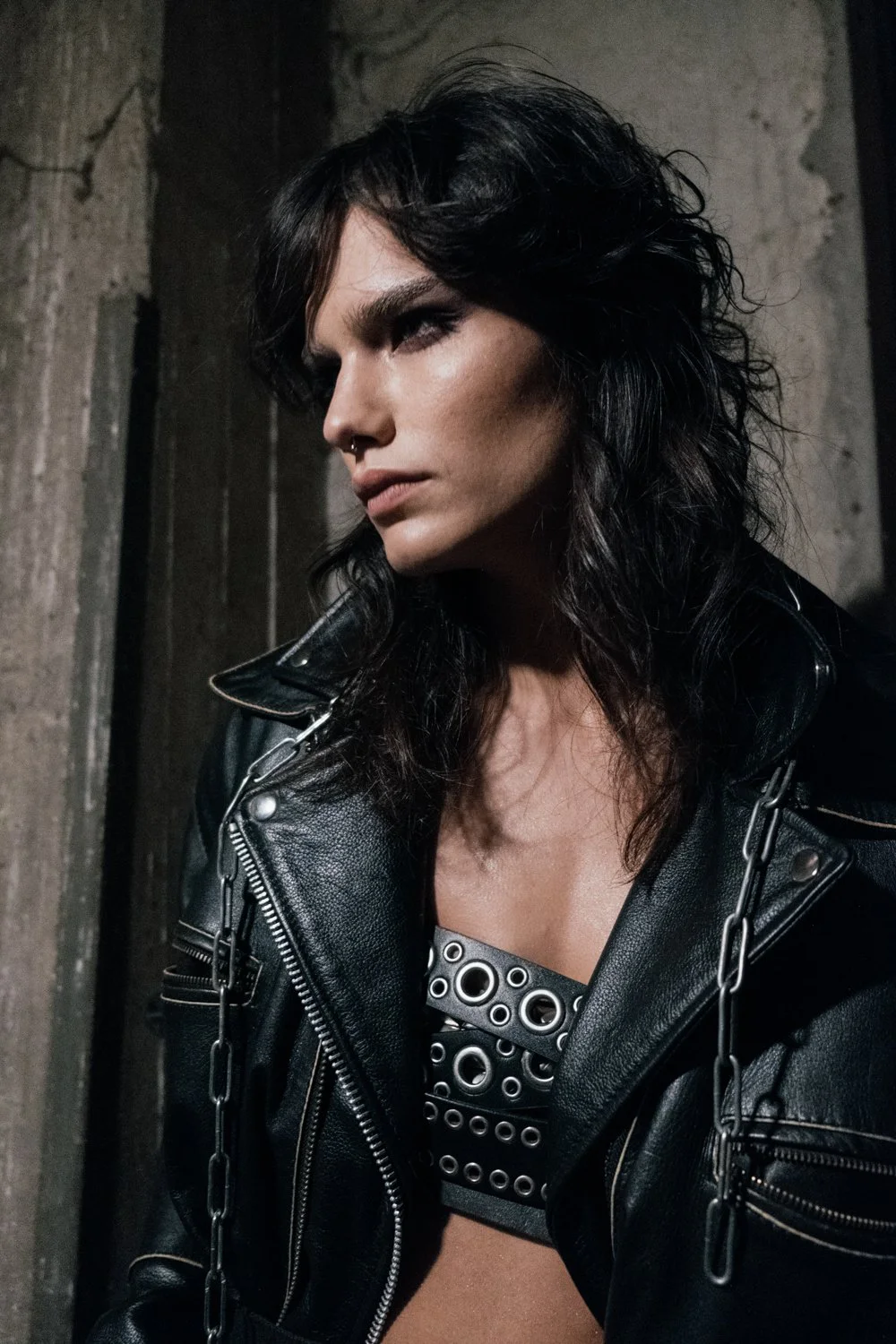 A woman with black, wavy hair wearing a black leather jacket and a studded black top, standing against a weathered wooden wall, with a serious expression.