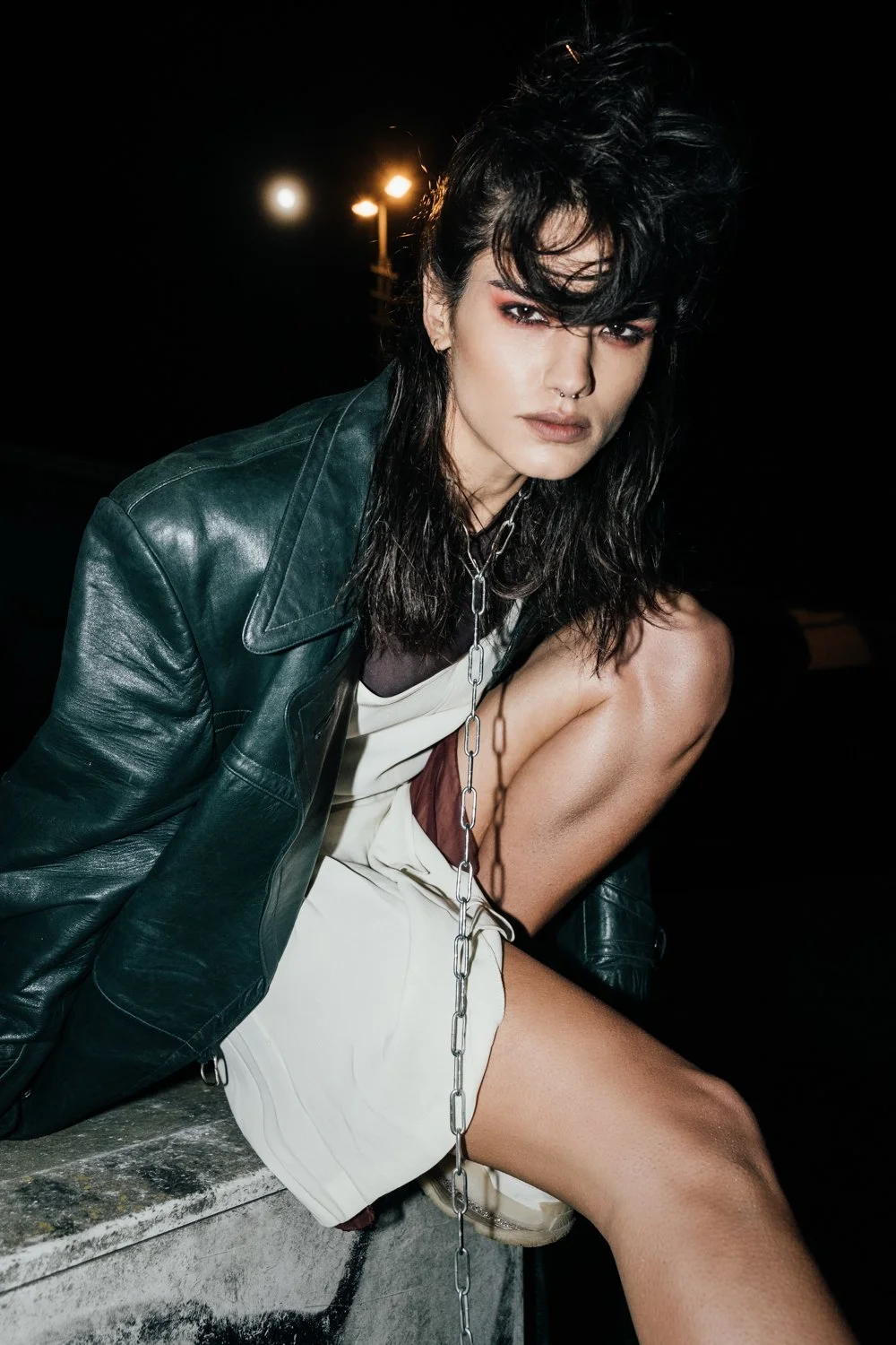 A woman with dark wavy hair, wearing a black leather jacket and white dress, sitting outdoors at night with a chain hanging from her neck, leaning against a surface, with streetlights in the background.