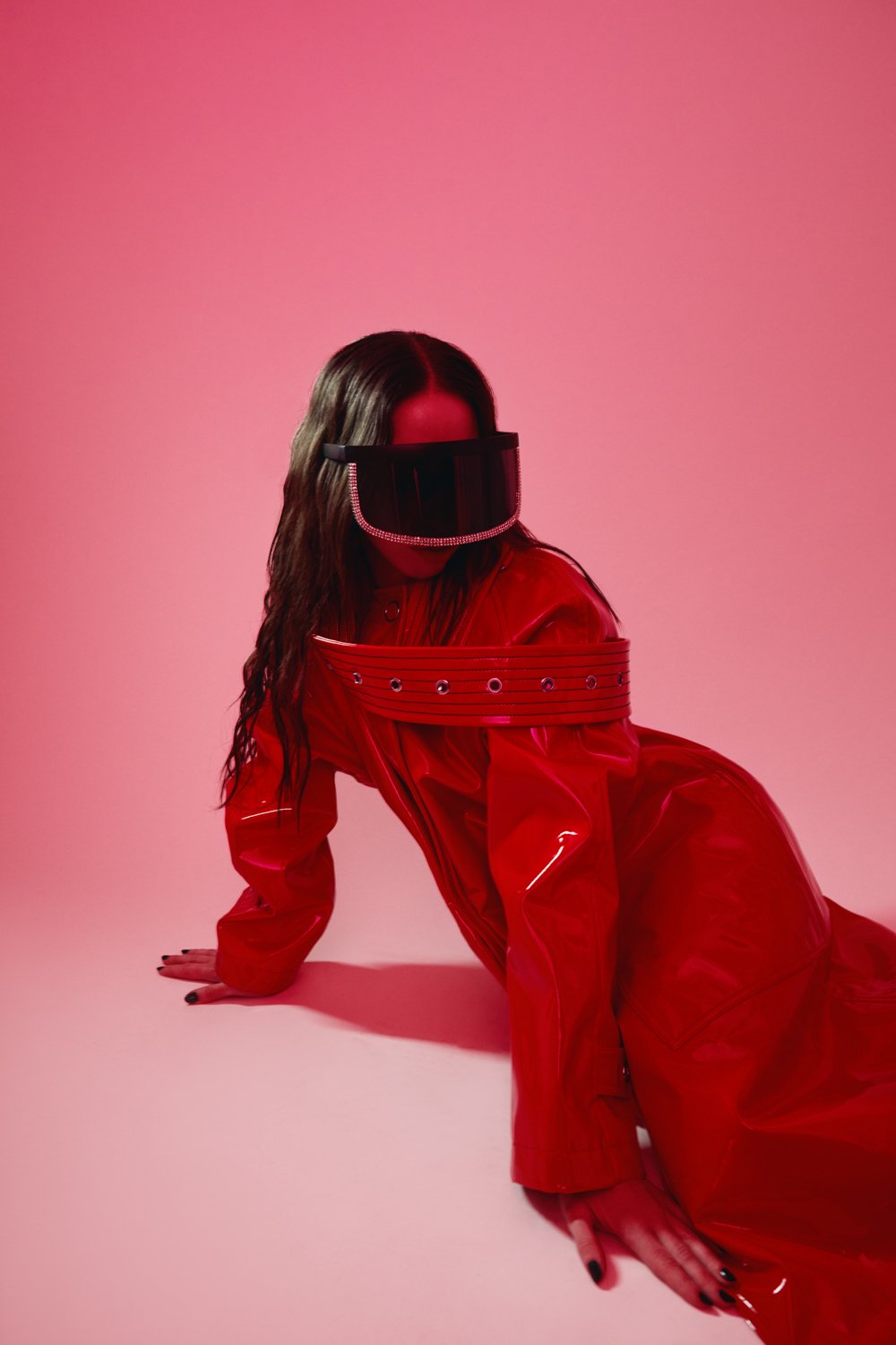 A woman in a red shiny outfit with a wide belt, wearing futuristic black visor glasses, and long wavy hair, on a pink background.