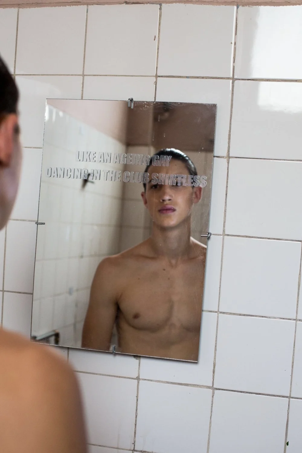 A shirtless man looking at his reflection in a bathroom mirror that has a quote on it. The mirror is mounted on a tiled wall and the quote says, 'LIKE AN AGENT'S GAY DANCING IN THE CLUB SHIRLESS'.