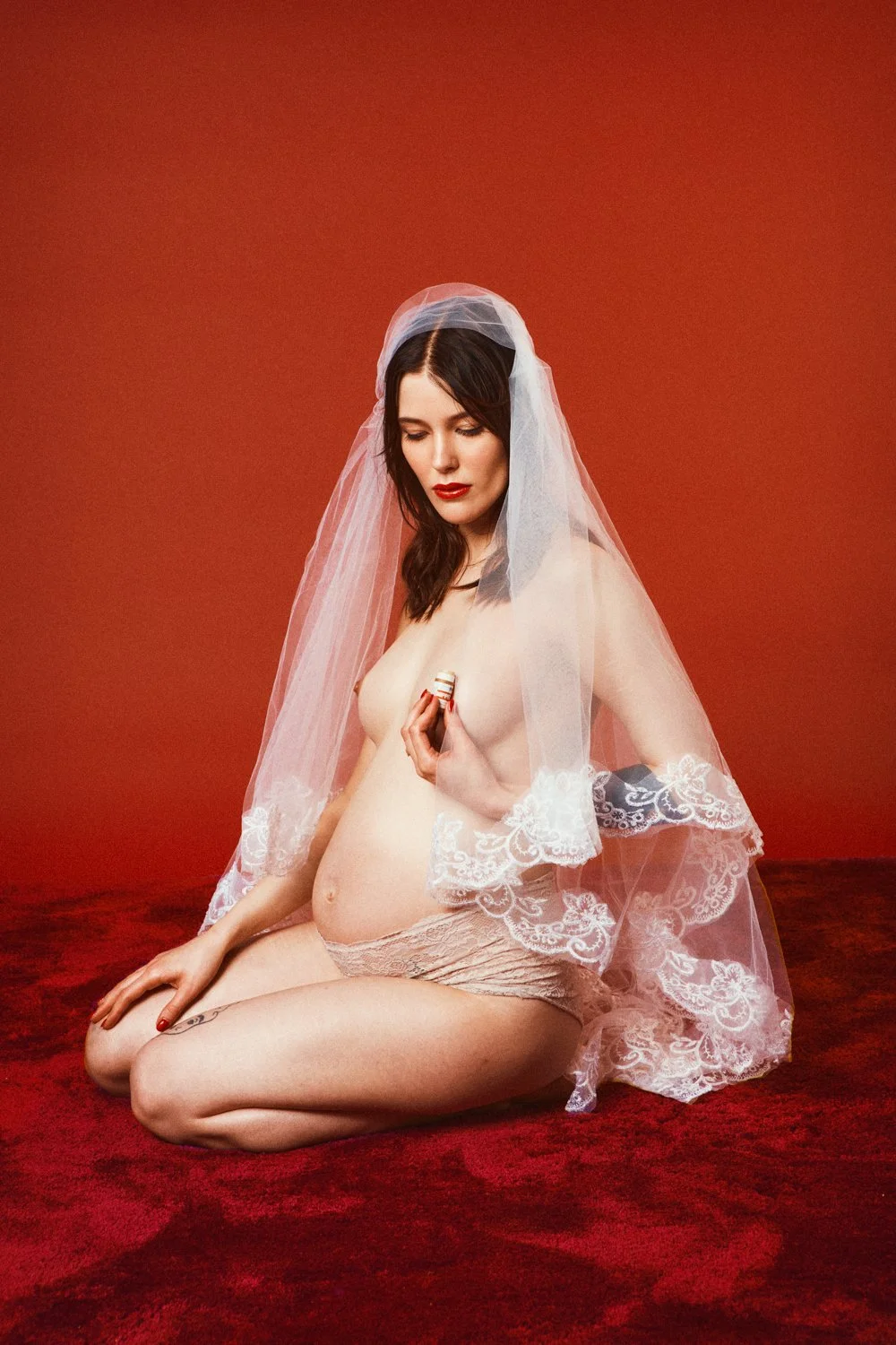 A woman with dark brown hair and red lipstick is kneeling on a red carpet, wearing only a lace shirt, shorts, and a sheer veil. She is holding a small object, possibly a ring, over her chest, with her eyes closed.