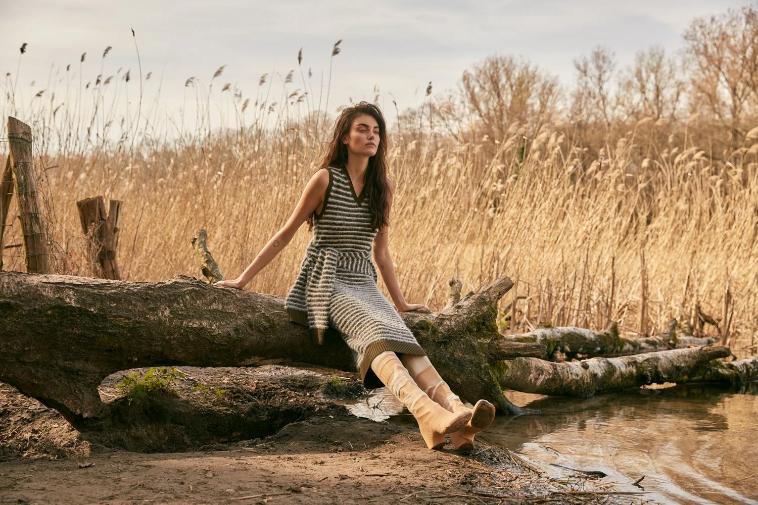 A young woman in a striped dress and beige boots sitting on a large log by a body of water, with tall dry grasses and trees in the background during what appears to be late afternoon or early evening.