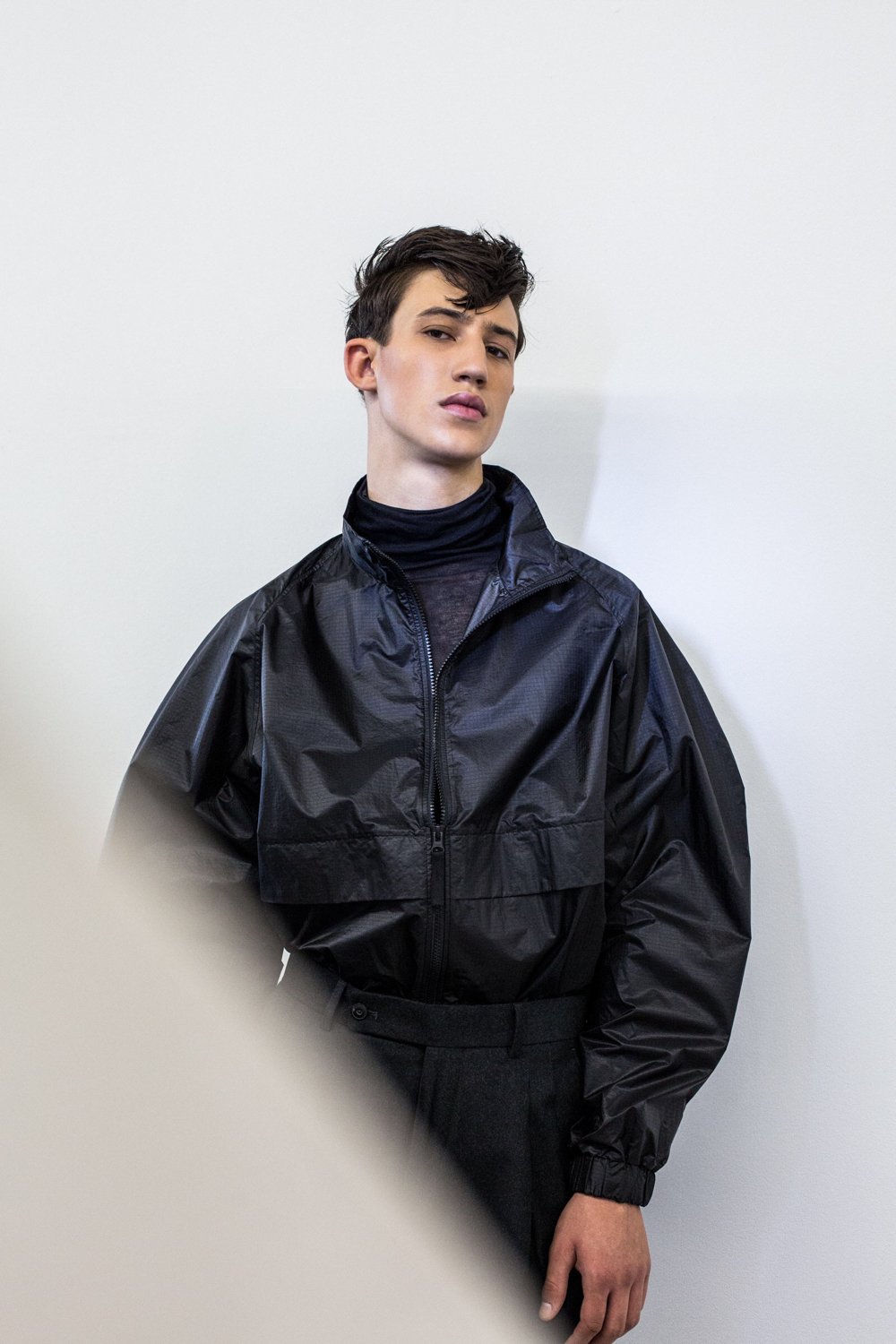 A young person with short dark hair wearing a black windbreaker jacket over a black turtleneck standing against a plain white wall.