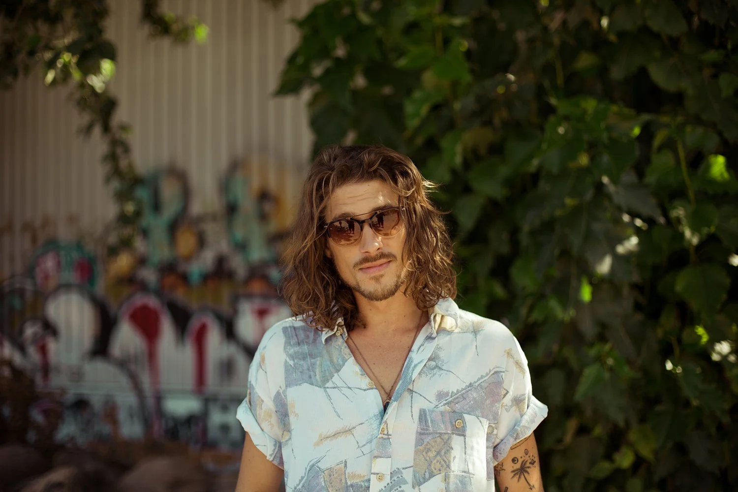 A young man with long, curly hair, wearing sunglasses and a patterned short-sleeve shirt, standing outdoors in front of a graffiti-covered wall and green foliage.