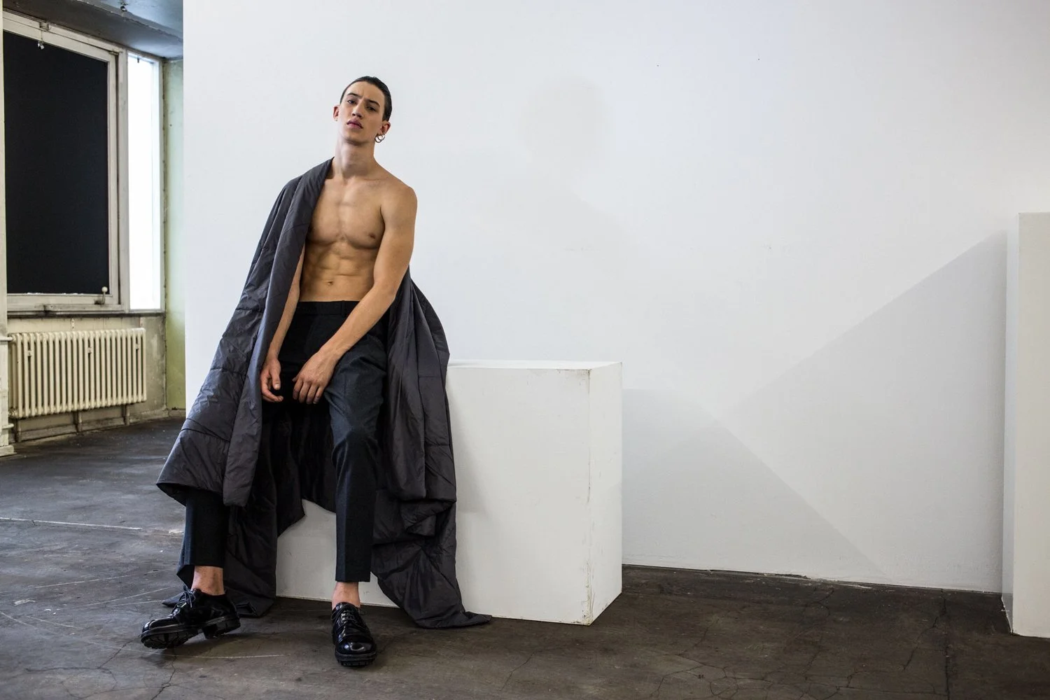 A shirtless man with a dark coat draped over his shoulders, sitting on a white pedestal in a minimalistic room with white walls, dark wooden floor, and a window with a radiator underneath.