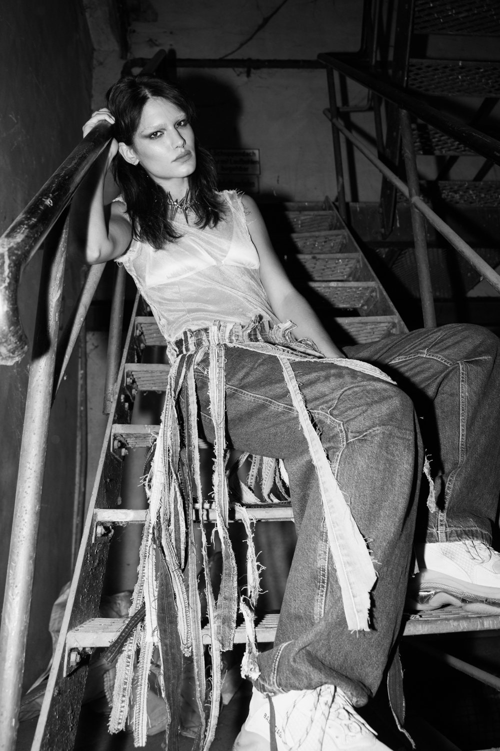 A young woman with dark hair and edgy makeup sitting on a metal staircase, wearing a sleeveless top and baggy jeans with hanging fabric strips.