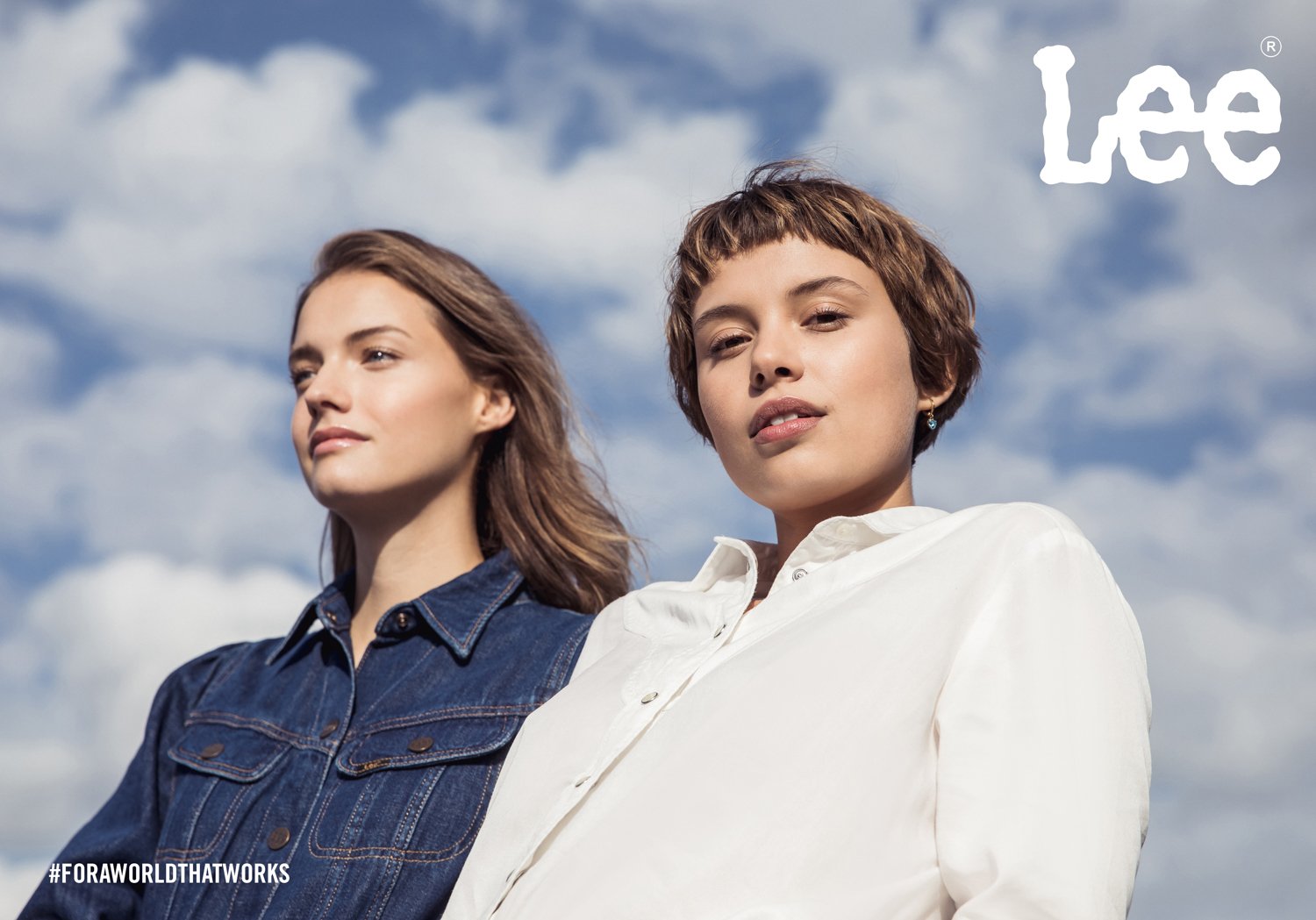 Two women standing outdoors under a cloudy sky, one wearing a denim jacket and the other in a white shirt, looking confidently at the camera.
