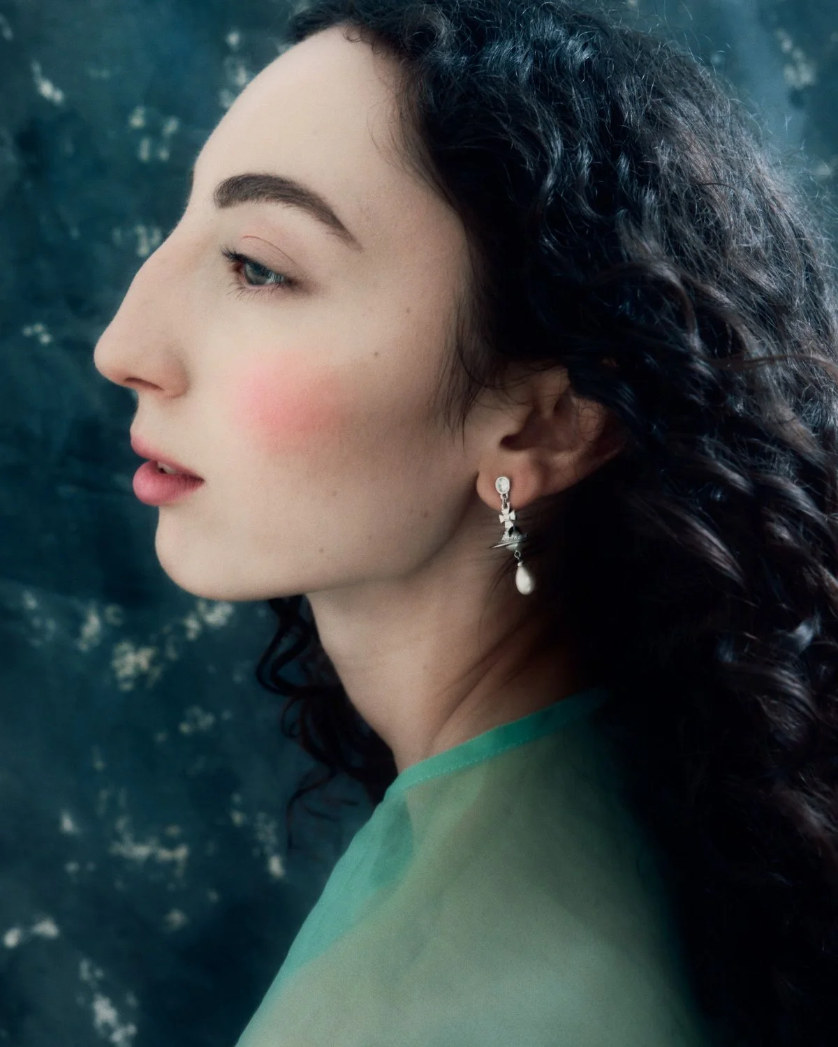 Side profile of a woman with dark, curly hair wearing earrings and a sheer green top, standing against a dark, textured background.