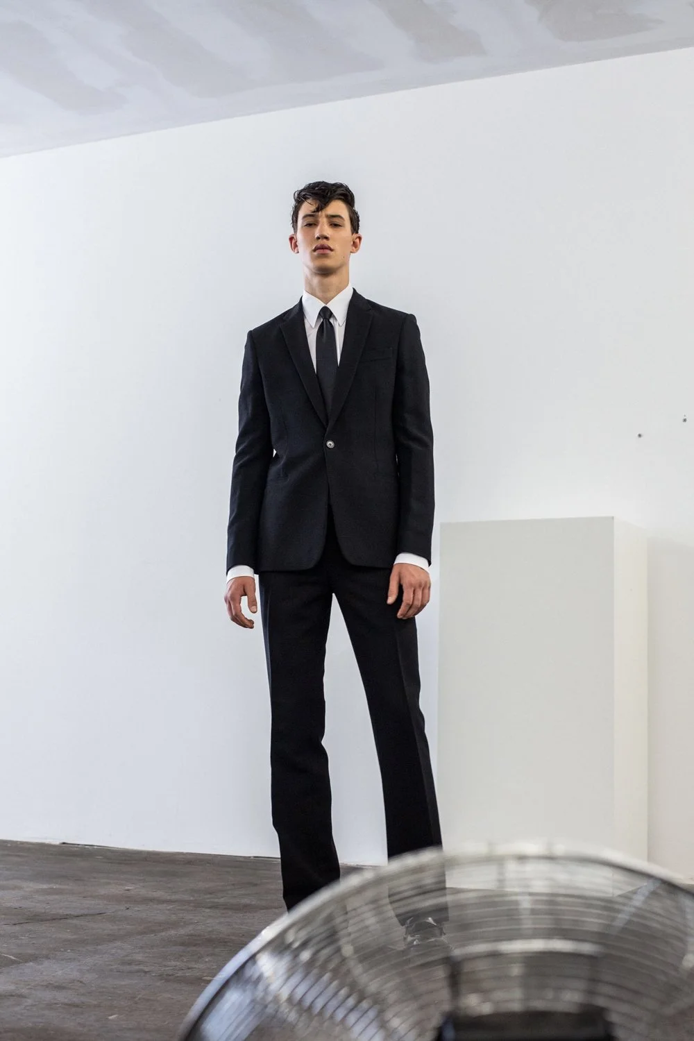 A young man in a black suit and tie standing indoors against a plain white wall.