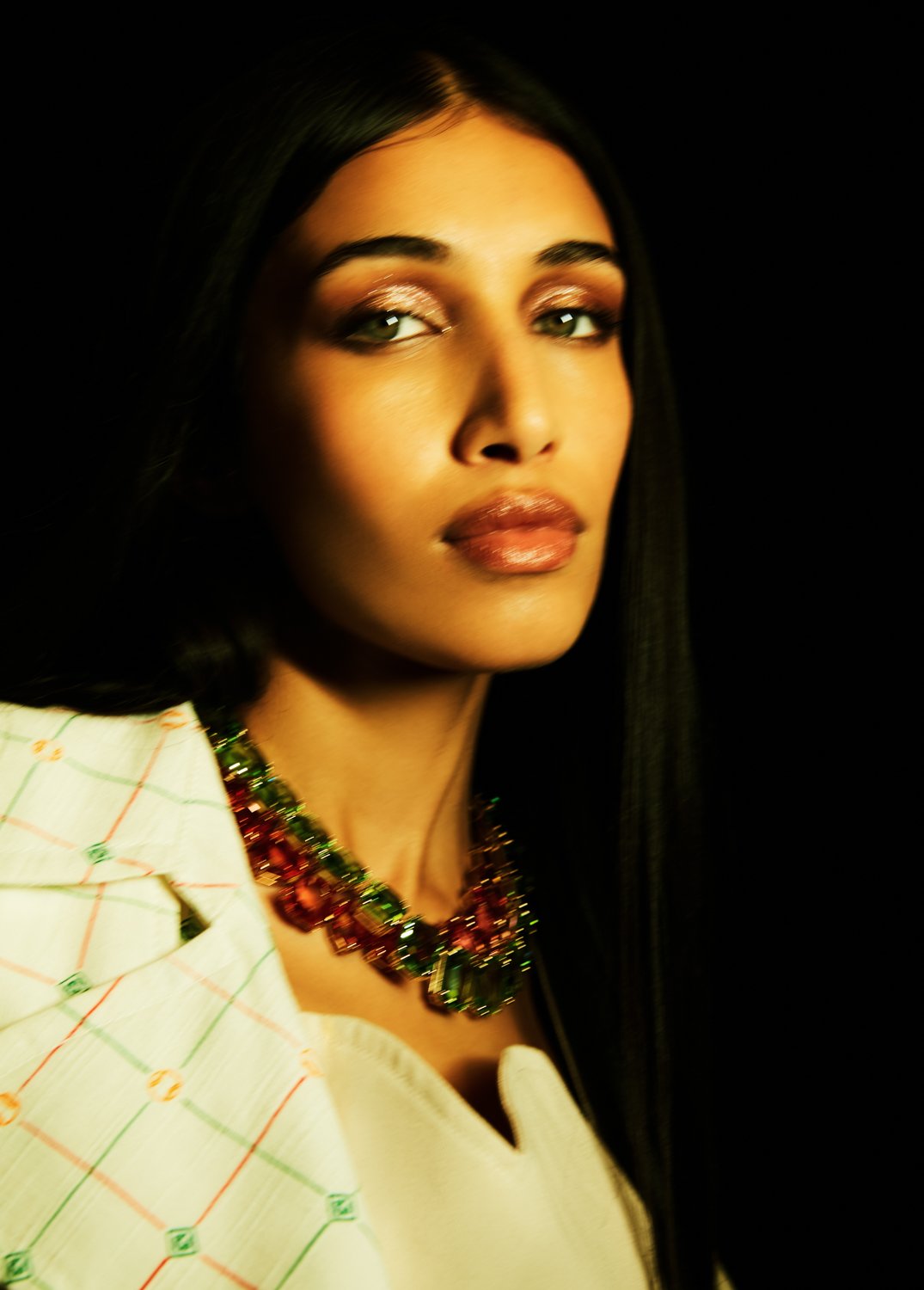 A woman with long black hair wearing a colorful necklace and a white patterned top against a black background.