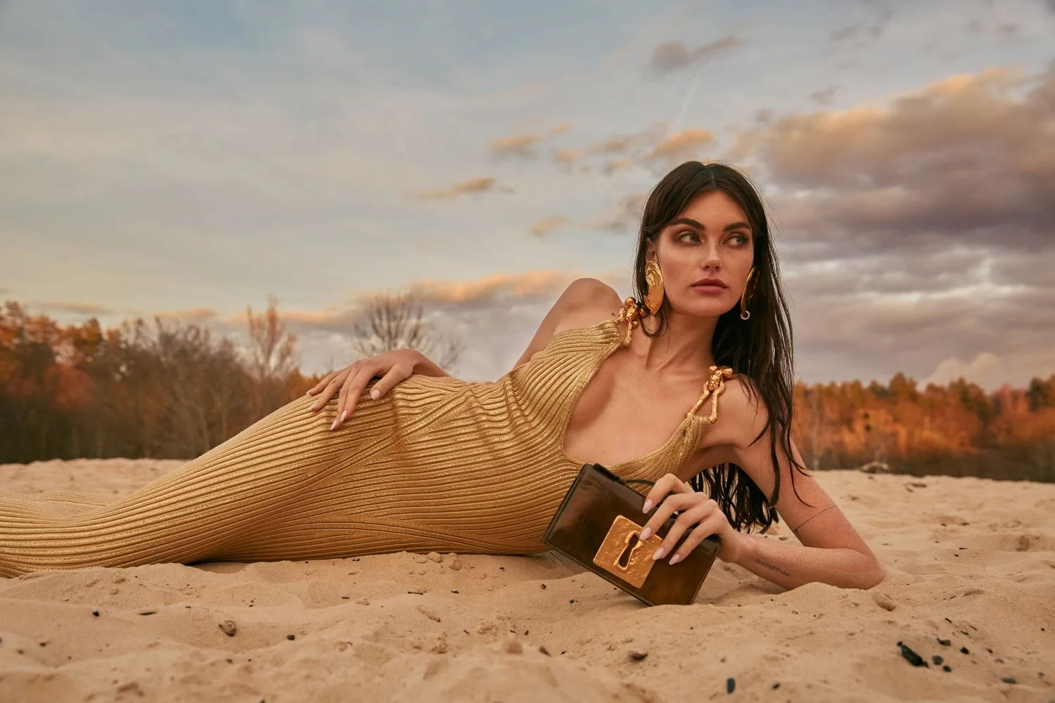 A woman lying on sandy ground outdoors during sunset, wearing a gold-colored dress with large gold earrings, holding a black clutch with a gold clasp, with a backdrop of trees with autumn foliage and a cloudy sky.