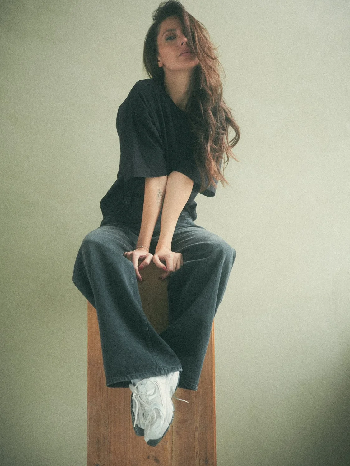 A woman with long wavy hair, sitting on a wooden block, wearing a black t-shirt, loose jeans, and white sneakers, against a plain light-colored wall.