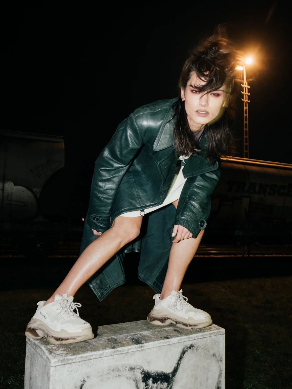 A woman with dark hair and makeup wearing a leather jacket, white sneakers, and black shorts, posing outdoors at night on a concrete block with streetlights in the background.