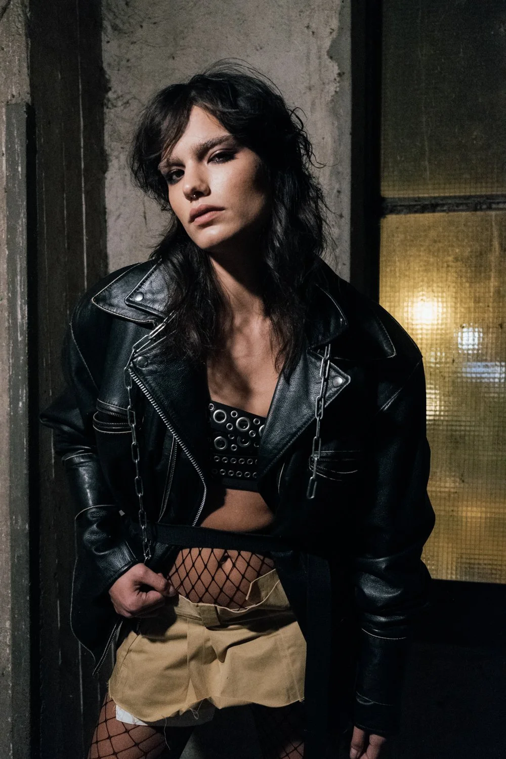A young woman with dark wavy hair, wearing a black leather jacket, black fishnet stockings, and beige shorts, standing in a dimly lit, industrial-style setting with concrete walls and a textured window in the background.