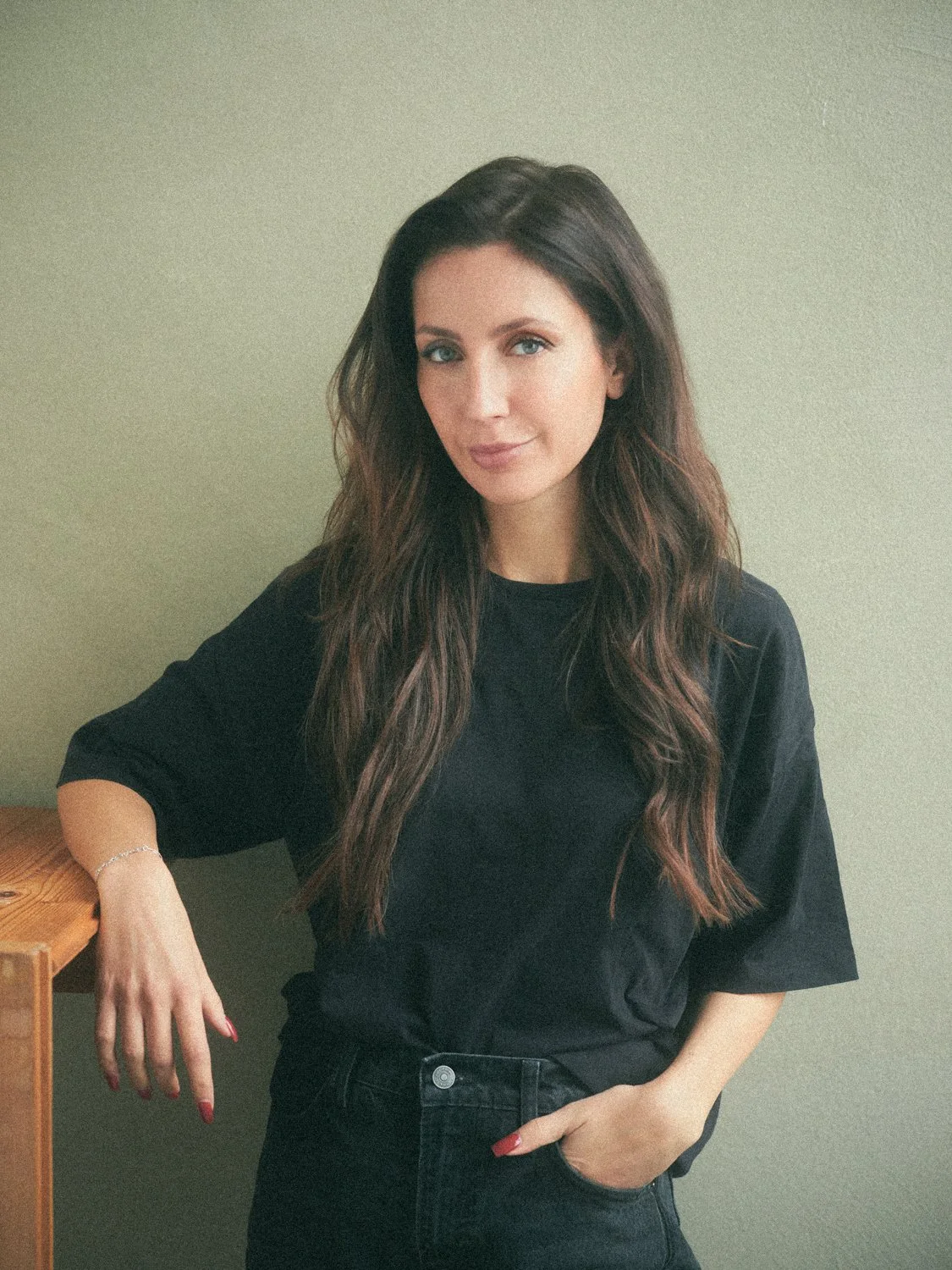 A woman with long, wavy brown hair and blue eyes, wearing a black t-shirt and black pants, standing against a plain, light-colored wall.