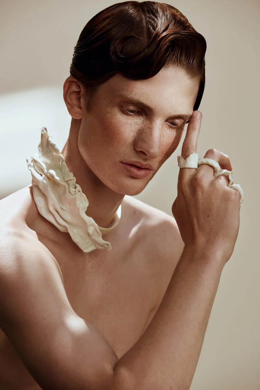 A person with short, wavy brown hair and freckles holding a finger to their temple. They have some white cream or lotion on their shoulder, neck, and fingers.