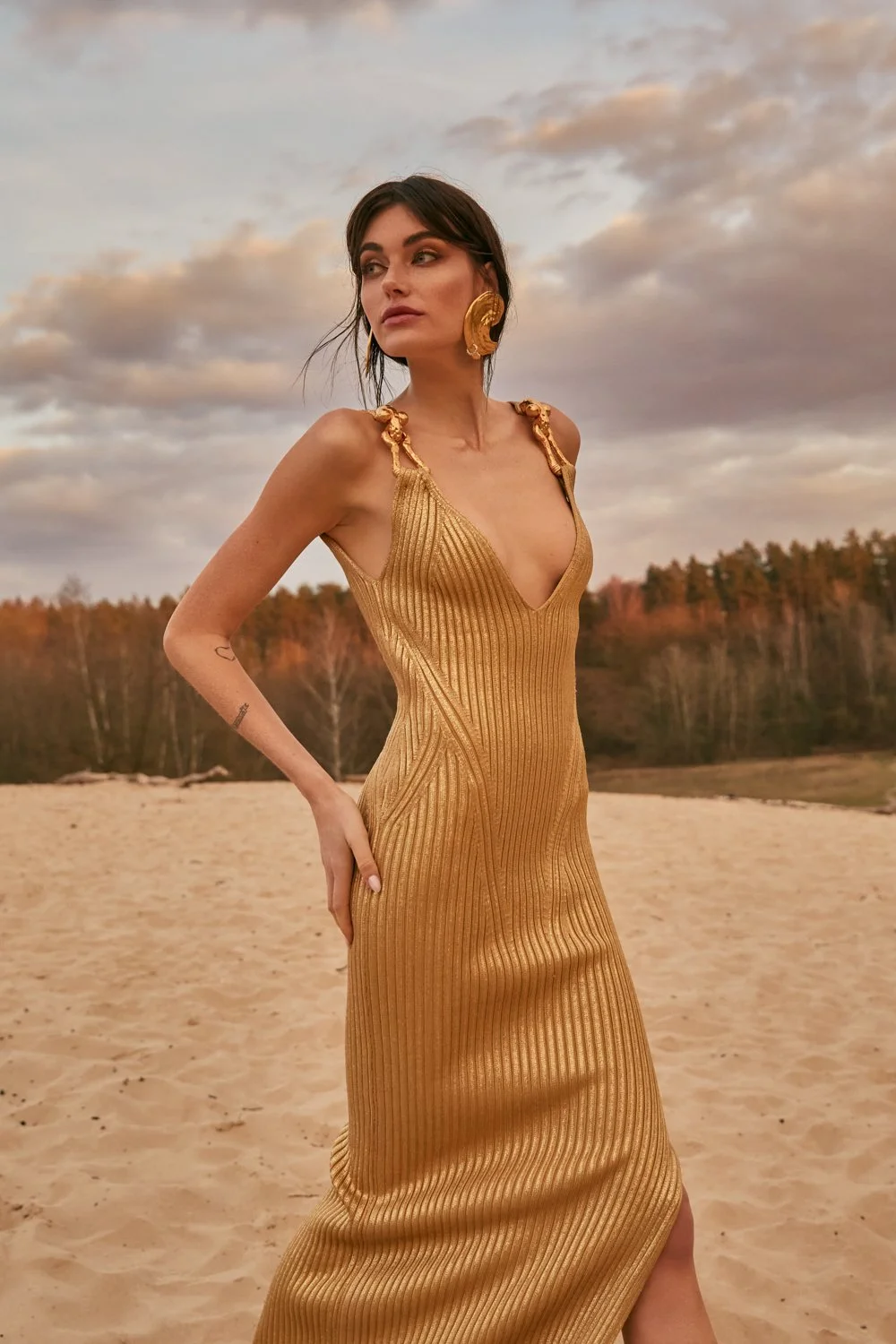 A woman in a gold dress standing on a sandy outdoor area with trees in the background and a cloudy sky overhead.