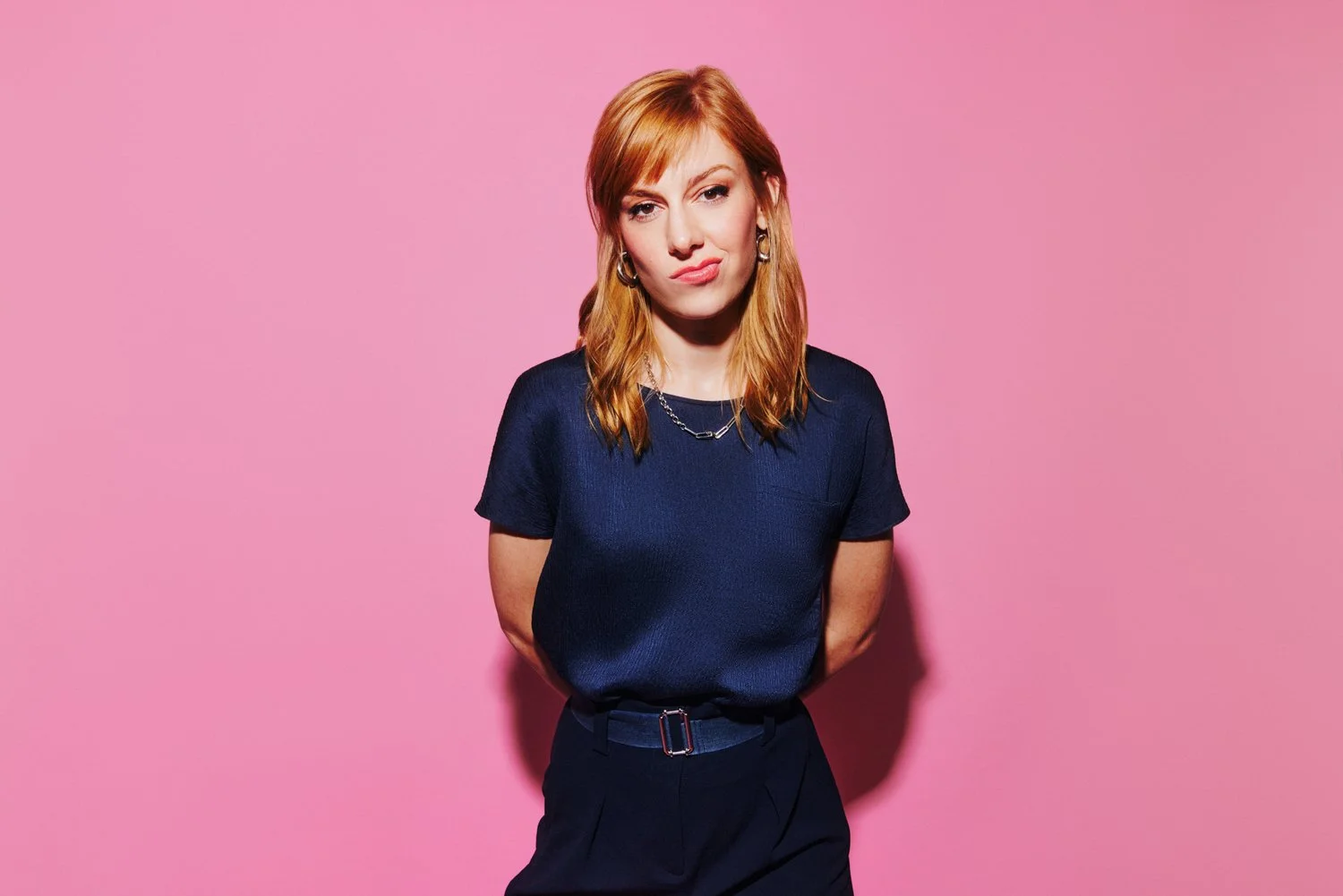 A young woman with red hair wearing a navy blue shirt and black pants, standing against a pink background, with a slightly puzzled expression.