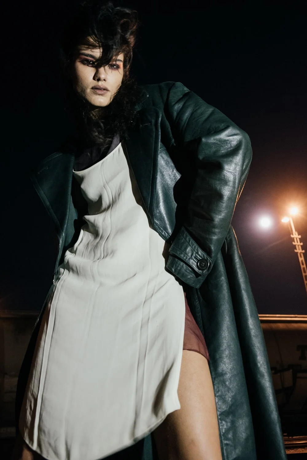 A young woman with dark, curly hair wearing a black leather jacket over a white dress standing outdoors at night under a bright streetlight.