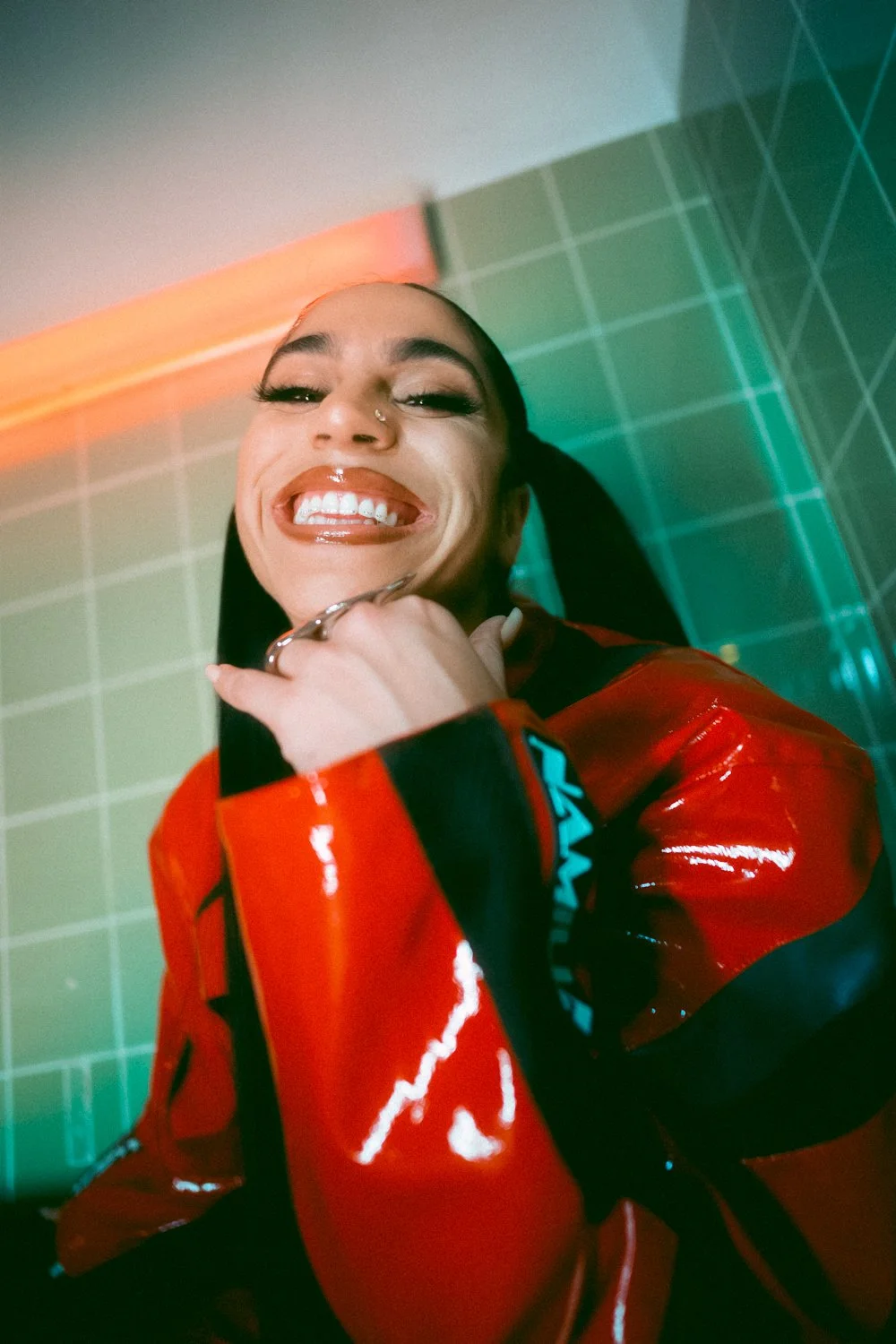 Close-up of a woman smiling and sticking her tongue out, wearing a red shiny jacket with black accents, against a background of green tiled wall with colored lighting.