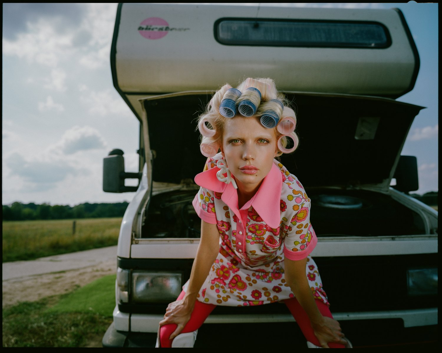 A woman with pink and blue hair rollers wearing a floral dress and pink collar, leaning forward with hands on her knees, in front of an open car hood in a rural setting under a cloudy sky.