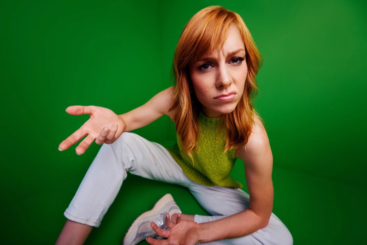 Woman with red hair in a green sleeveless top and white pants, sitting on the floor in front of a green background, making a hand gesture with a serious expression.