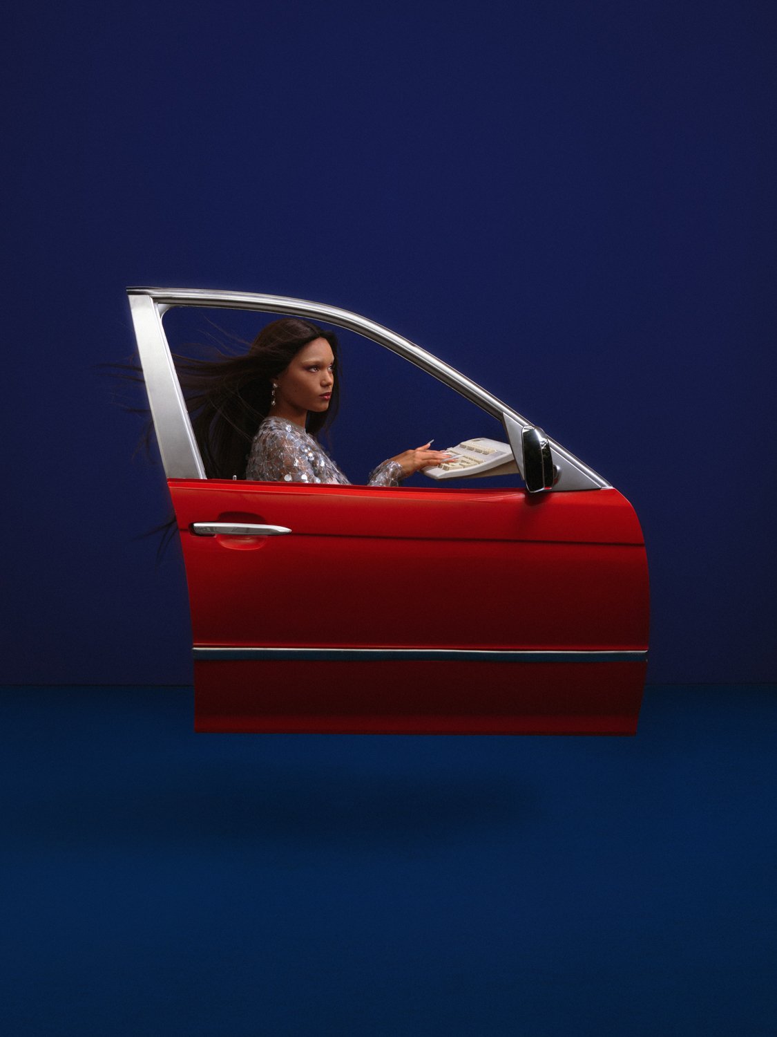 A woman sitting inside a red car, holding a telephone receiver, with a dark blue background.