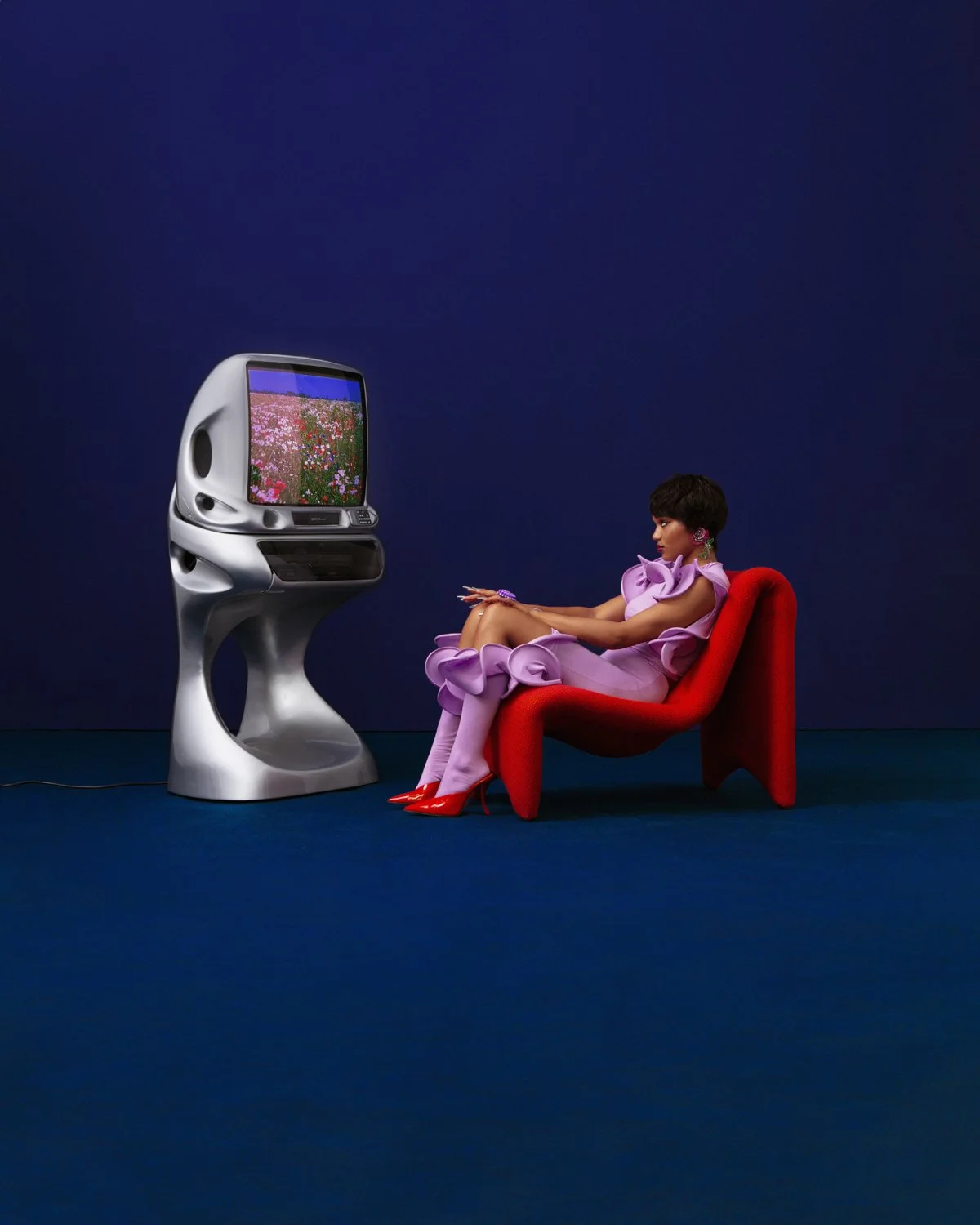 A woman in a pink dress and red high heels sitting on a red chair, looking at a vintage silver television displaying a field of pink and red flowers. The background is dark blue.