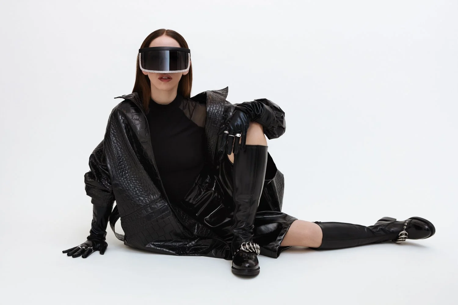 A woman dressed in all black with a futuristic visor, sitting on the floor against a plain white background, wearing a black leather jacket, gloves, thigh-high boots, and sunglasses.