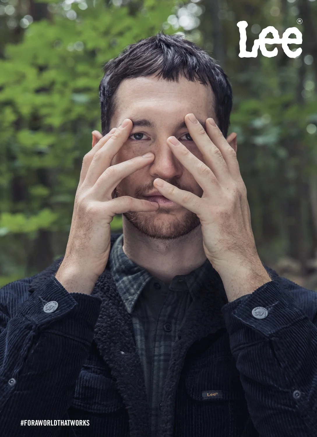 A man with short dark hair and a beard, holding his hands in front of his face, outdoors in a forested area.