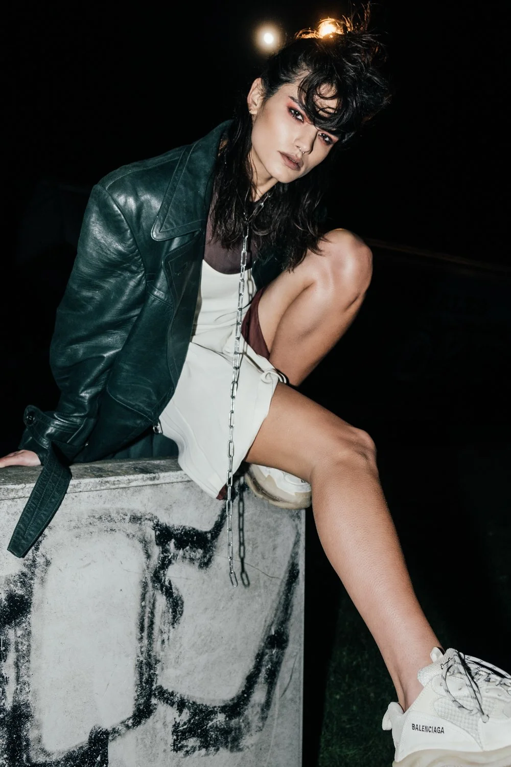 A woman with dark, curly hair and makeup, wearing a leather jacket, shorts, and sneakers, sitting on a concrete structure at night with a chain hanging from her neck.