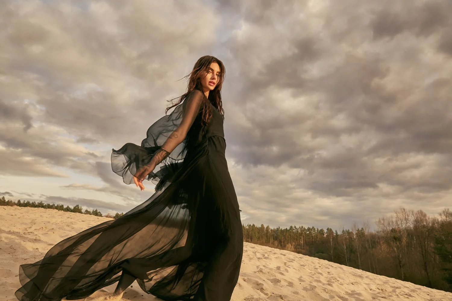 A woman dressed in black flowing gown standing on a sandy area under a cloudy sky, with trees in the background.
