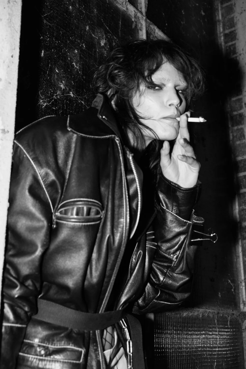 A woman with tousled dark hair, wearing a leather jacket, smokes a cigarette, leaning against a brick wall.