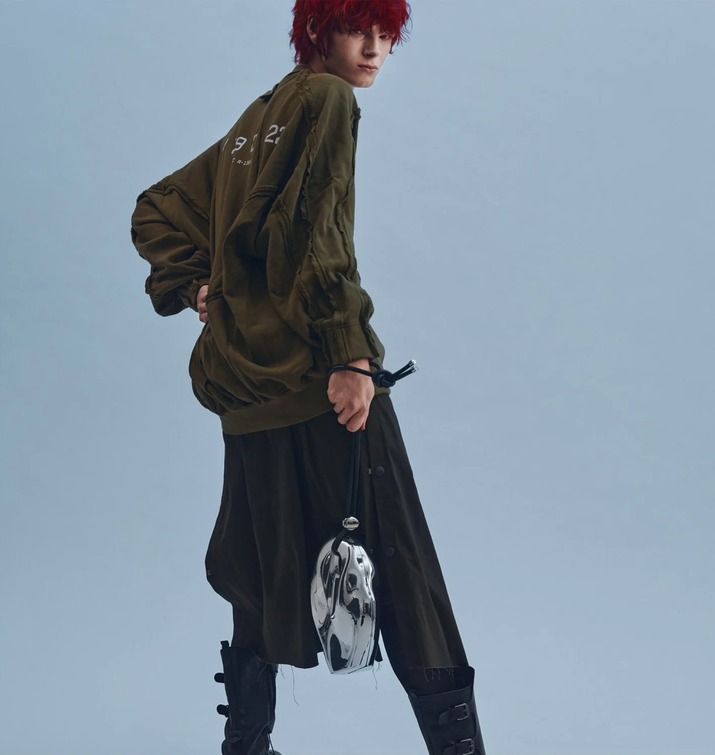 A young person with red curly hair wearing a dark green bomber jacket and black pants. They are holding a shiny silver helmet in their right hand and standing against a plain light blue background.