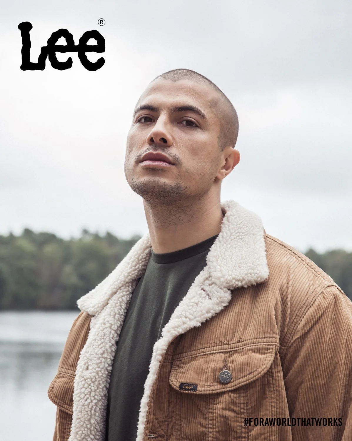 Portrait of a young man wearing a brown jacket with a shearling collar, standing outdoors by a lake with trees in the background.