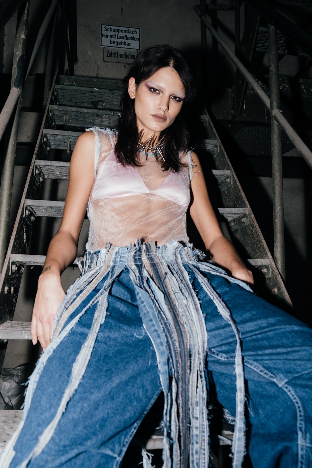 A young woman with black hair, dark makeup, and a spiked necklace, sitting on a metal staircase in an industrial setting.