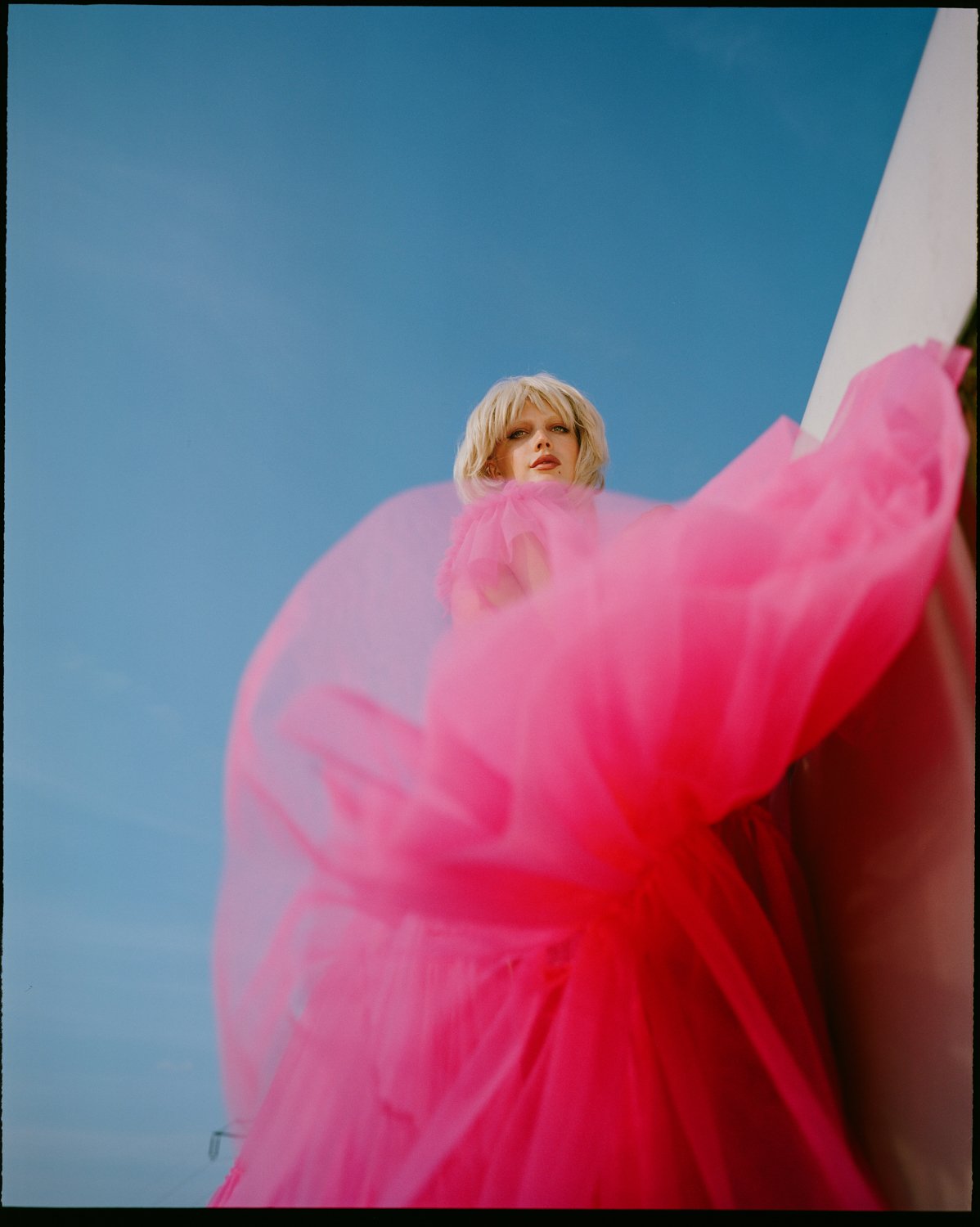 A woman with blonde hair in a pink dress stands against a blue sky, seen from below with flowing pink fabric.