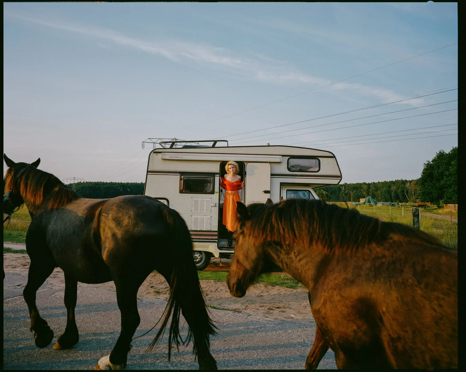 A woman in an orange dress standing in the doorway of a white recreational vehicle, with two horses grazing nearby on an open field at sunset.