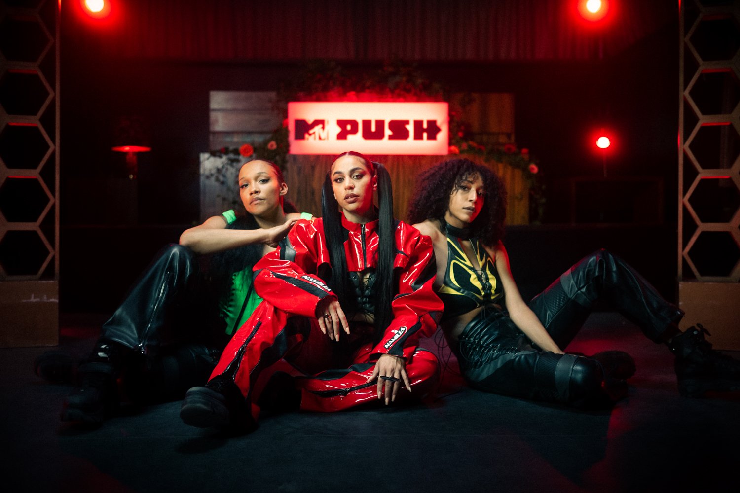 Three women sitting on the floor in front of an 'MTV PUSH' sign, with a dark background and red lighting.